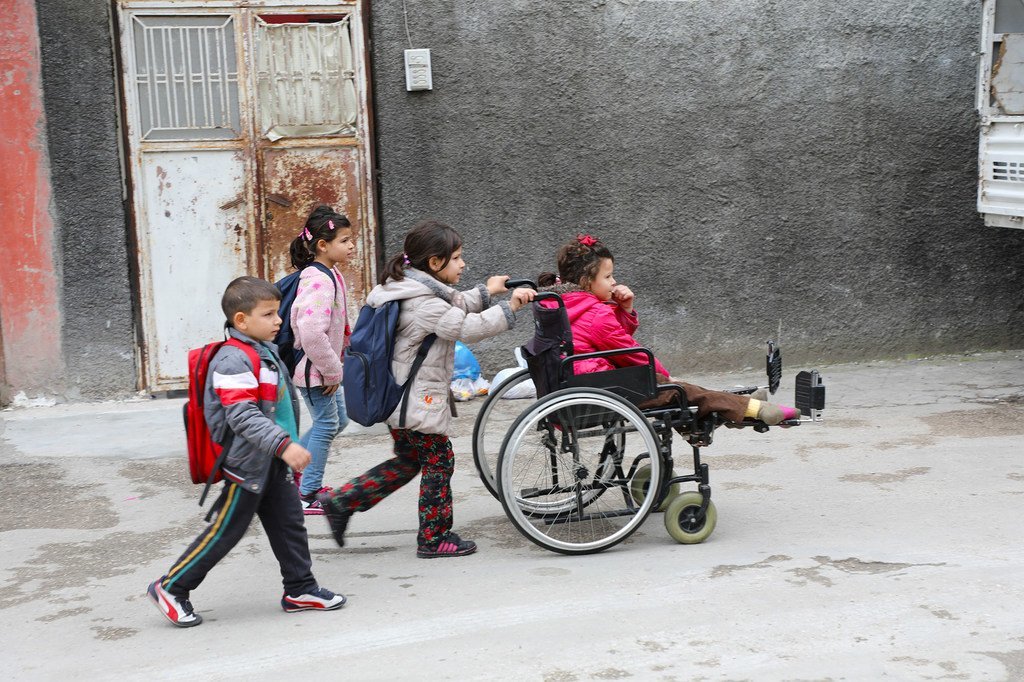 طفلة لاجئة من ذوي الإعاقة تجلس على كرسي متحرك تساعدها أختها بينما تتوجه هي وأشقاؤها إلى المدرسة في أضنة، تركيا.