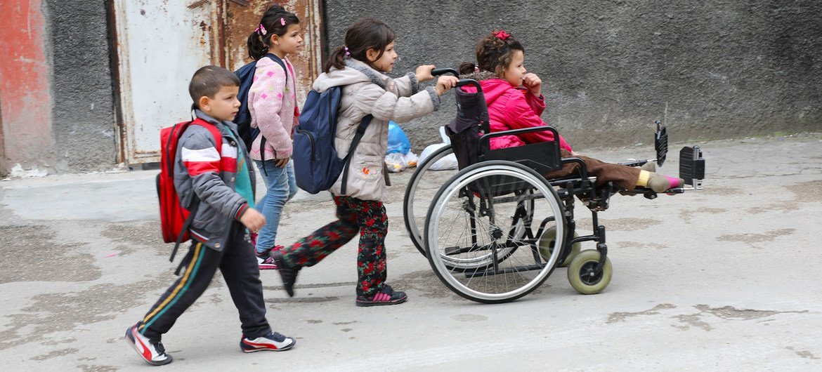 طفلة لاجئة من ذوي الإعاقة تجلس على كرسي متحرك تساعدها أختها بينما تتوجه هي وأشقاؤها إلى المدرسة في أضنة، تركيا.
