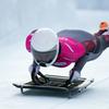 Uma atleta de luge, vestida com um traje rosa e cinza, desce em alta velocidade uma pista de gelo durante um evento de esportes de inverno.