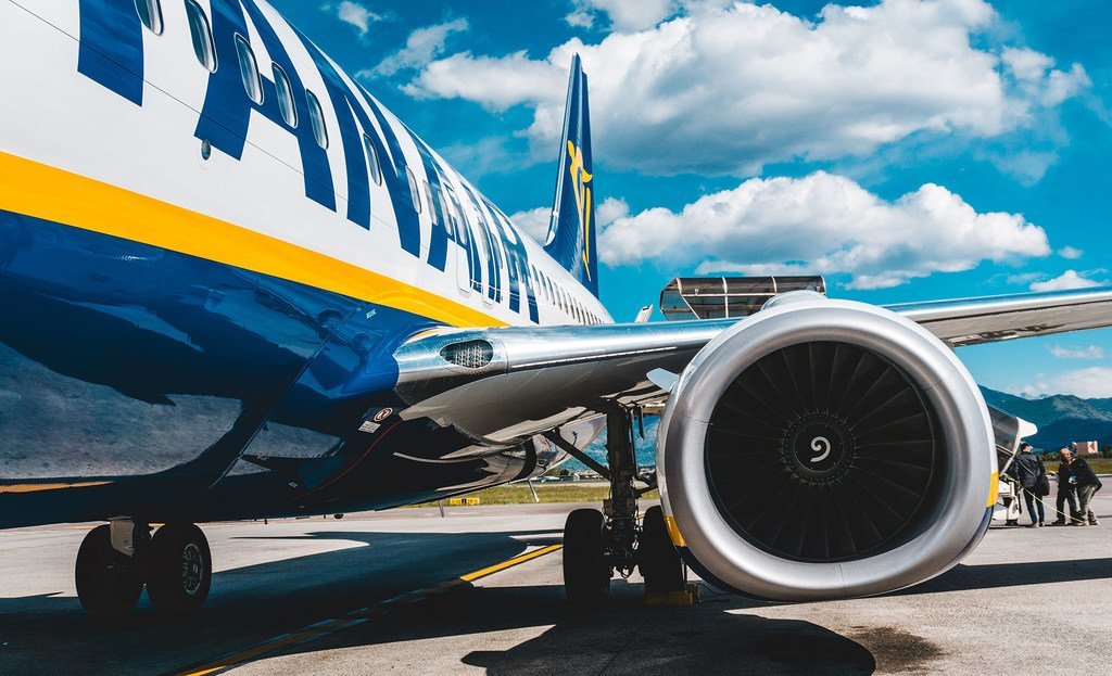 Primer plano de un avión de Ryanair en la pista, donde se aprecian su gran motor a reacción y el tren de aterrizaje contra un cielo azul con nubes.