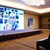 تستمع امرأة ترتدي بدلة سوداء وقناعاً للوجه إلى عرض تقديمي افتراضي على شاشة كبيرة. تعرض الشاشة ممثلة UNICEF، إيتي هيغينز، وهي امرأة محجبة، تتحدث إلى الحضور عن الأزمة اللبنانية وتأثيرها على تعليم الشباب.