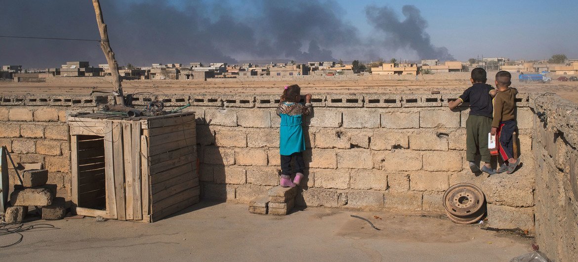 In Iraq, children look over a wall at clouds of smoke from burning oil wells, the result of oil fires set by ISIL.