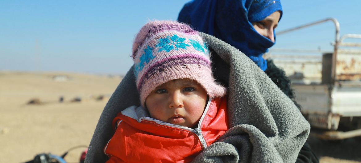 सीरिया के पूर्वी ग्रामीण इलाक़े - रक़्क़ा की एक अनौपचारिक बस्ती में, एक महिला अपने बच्चे के साथ, सर्दियों के कपड़े लेने के इन्तेज़ार में.