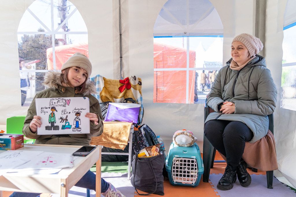 A nine-year-old Ukrainian girl holds a drawing of her family while sitting in a learning center with her mother and cat (in a blue basket) in Romania.