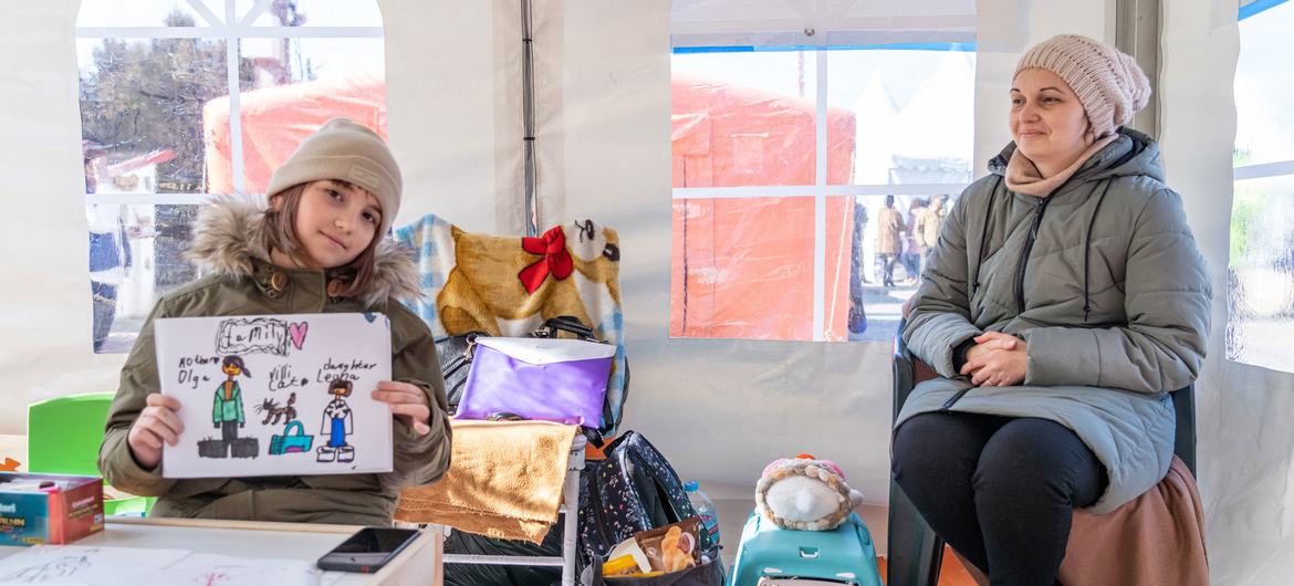 A nine-year-old Ukrainian girl holds a drawing of her family as she sits in a learning hub with her mohther and cat (in blue basket) in Romania.