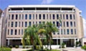 A large, modern multi-story office building with a glass and concrete facade is shown from the street level, surrounded by palm trees and other landscaping.
