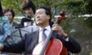 An Asian man in a suit plays a cello on a stage with other musicians in the background.