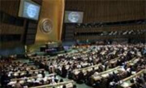 Wide shot of the General Assembly Hall at the United Nations in New York, showing numerous delegates seated at desks in a large, wood-paneled circular room.