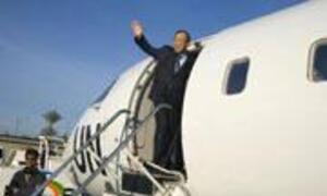 A pilot in uniform and a scarf stands on the stairs of a white airplane, waving goodbye.