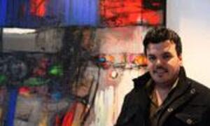 A man in a dark jacket stands smiling in front of a colorful abstract painting in an art gallery.