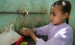 A young girl stands at a water pump, drinking water from a green container.
