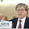 A man with grey hair and glasses wearing a suit and tie, speaking at a formal meeting with a 'BELARUS' placard in front of him.