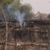 A burnt hut with smoke rising from the wreckage, located in a rural village setting.