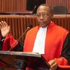 A male judge in a red and black robe speaks at a podium in a courtroom.
