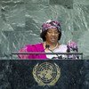 A woman in a floral headwrap and formal attire speaks at a podium featuring the United Nations emblem.