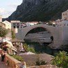 A panoramic view of the historic Stari Most (Old Bridge) spanning the Neretva River in Mostar, Bosnia and Herzegovina, with tourists walking along the adjacent stone-paved street.