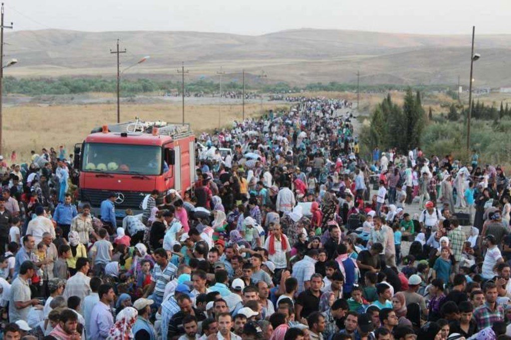 Uma grande multidão de refugiados sírios atravessa uma fronteira, com um caminhão de bombeiros vermelho em primeiro plano.