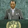 Un homme en costume à fines rayures et lunettes se tient à un podium orné du logo des Nations Unies , parlant dans des microphones devant un mur de marbre vert.