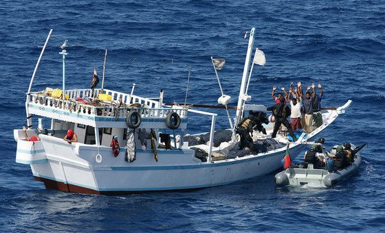 Un bateau de pêche blanc est abordé par des forces spéciales armées à bord d'un bateau pneumatique, plusieurs hommes à bord du bateau de pêche levant les mains.