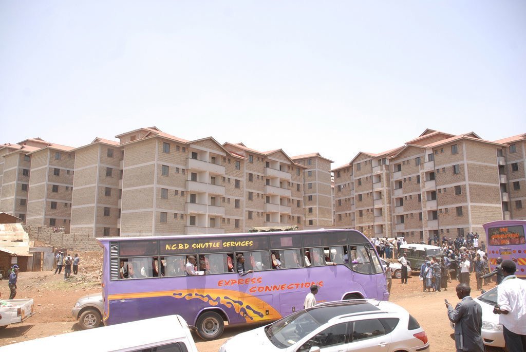 Basi la usafiri la rangi ya zambarau lenye maandishi 'NCBD SHUTTLE SERVICES' na 'EXPRESS CONNECTIONS' limeegeshwa mbele ya jengo kubwa la makazi la ghorofa nyingi linalojengwa. Umati wa watu na magari kadhaa wamekusanyika kwenye eneo la udongo.