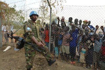 A peacekeeper stands guard at UN House while displaced children look on behind a fence.