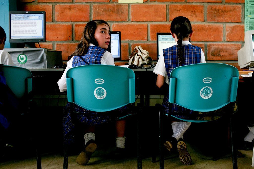 Dos estudiantes, todas mujeres y vestidas con uniforme escolar, están sentadas frente a una computadora en un aula con una pared de ladrillos.