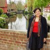 Une femme souriante, vêtue d'un haut rouge et d'un manteau sombre, se tient sur un pont de briques dans un parc, avec un canal et des maisons traditionnelles en arrière-plan.