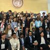 A large group of young people in formal and business attire pose for a group photo at the Global Youth Forum, with the United Nations emblem on the wall behind them.