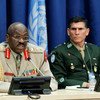 Two military generals sit at a conference table with a United Nations flag in the background.