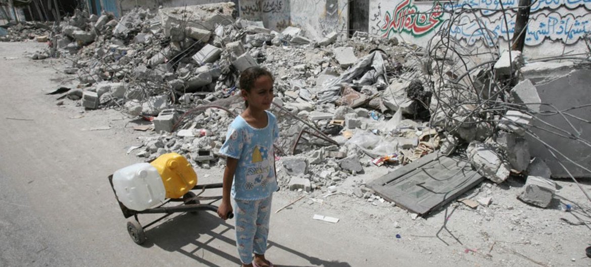 On 19 July 2014 in the State of Palestine, passing the rubble of homes destroyed in an Israeli air strike, a girl uses a hand truck to transport jerrycans filled with water, in the town of Rafah in southern Gaza.