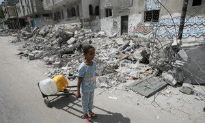On 19 July 2014 in the State of Palestine, passing the rubble of homes destroyed in an Israeli air strike, a girl uses a hand truck to transport jerrycans filled with water, in the town of Rafah in southern Gaza.