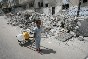 On 19 July 2014 in the State of Palestine, passing the rubble of homes destroyed in an Israeli air strike, a girl uses a hand truck to transport jerrycans filled with water, in the town of Rafah in southern Gaza.