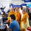 Trois moines bouddhistes vêtus de robes orange se tiennent dans un magasin d'électronique, observant un ordinateur portable présenté par un client.