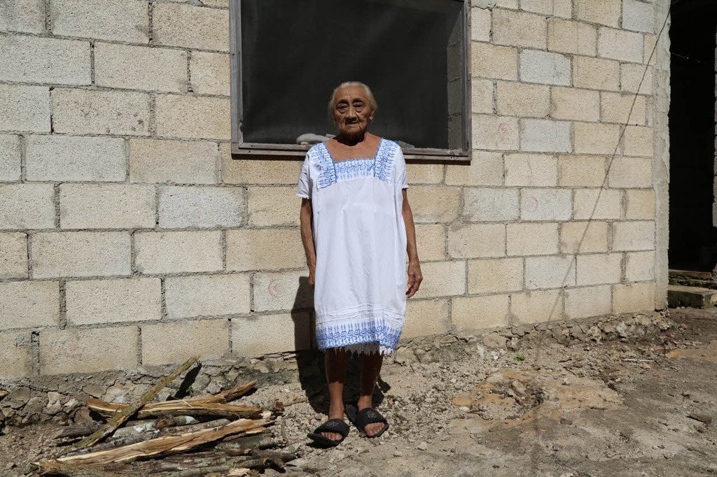 Una anciana con un vestido blanco bordado se encuentra de pie frente a un edificio de bloques de cemento sin terminar.
