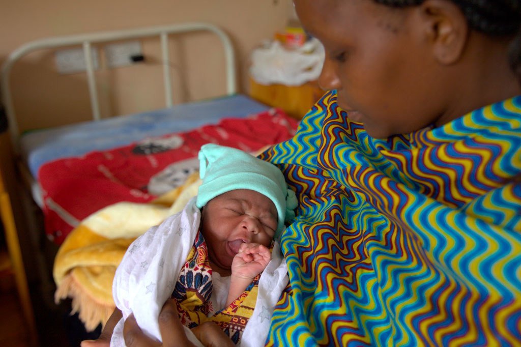 Mama akiwa amembeba mtoto wake mchanga katika kituo cha afya nchini Malawi. Mtoto, akiwa amefunikwa na blanketi na amevaa kofia, analia huku mama akiitazama chini.