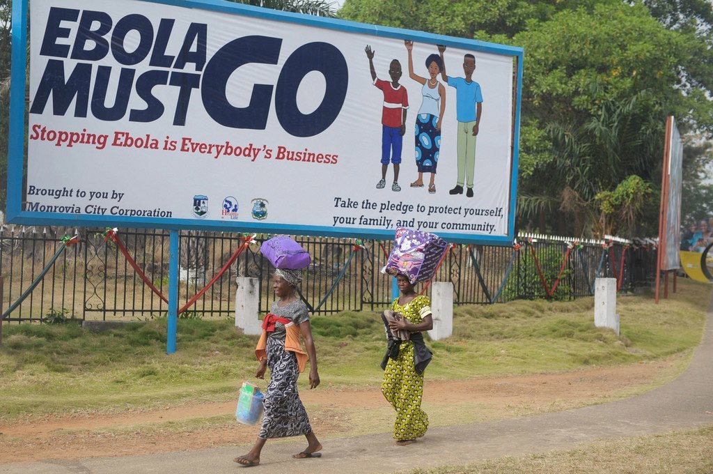 Um cartaz em Monróvia, Libéria, com a mensagem "O EBOLA DEVE IR EMBORA" e ilustrações de pessoas, enquanto duas mulheres passam caminhando por uma estrada de terra.