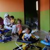 An Omani woman sits on a sofa holding a baby while several other children play with baby walkers in a colorful room.