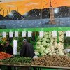 Vendors at a vegetable market stall in Syria, with a large mural of a city skyline on the wall behind them.