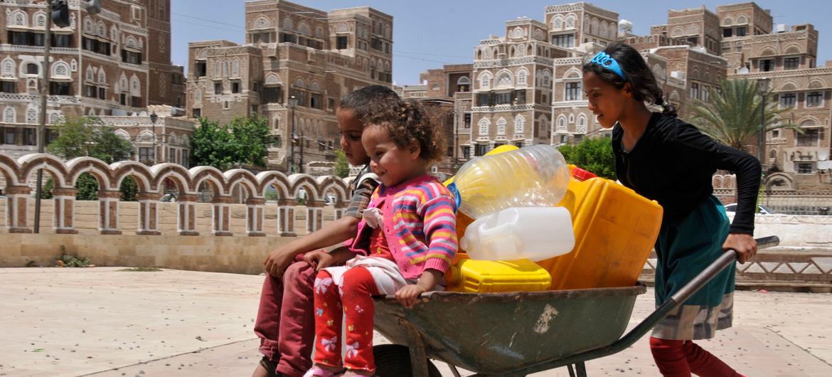 A girl pushes two younger children  in a wheelbarrow that also bears several jerrycans in Sana’a, the capital of Yemen.