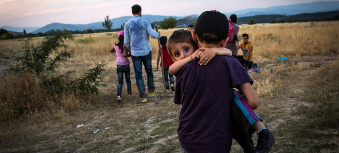 一名叙利亚难民青年抱着自己的弟弟跨过希腊和前南马其顿共和国边境。难民署图片/A. McConnell