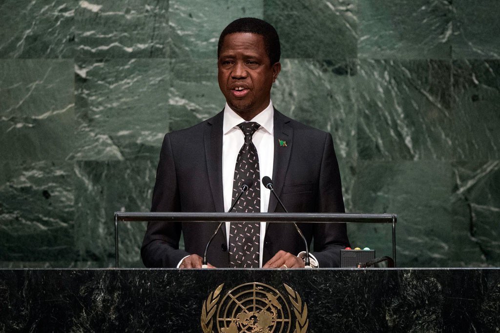 Mwanamume aliyevaa suti amesimama kwenye jukwaa lenye nembo ya UN , akizungumza katika hafla rasmi katika ukumbi wenye ukuta wa marumaru ya kijani.