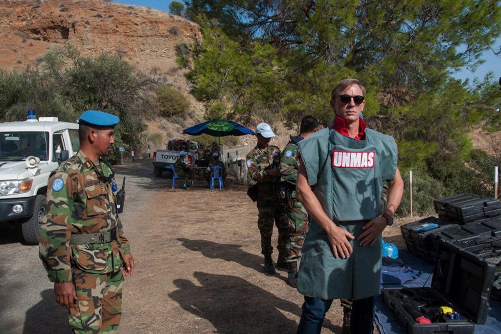 Дэниел Крейг в защитном костюме UNMAS стоит рядом с военнослужащими на открытом воздухе.