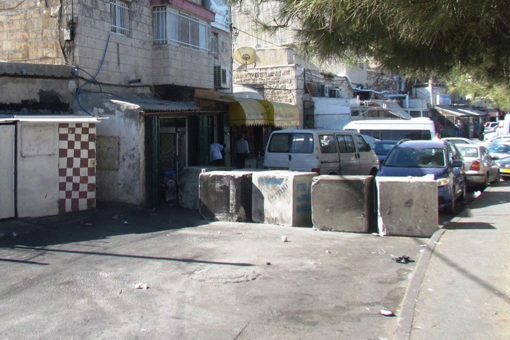 مشهد شارع في القدس يضم مباني حجرية وحواجز خرسانية وسيارات متوقفة.