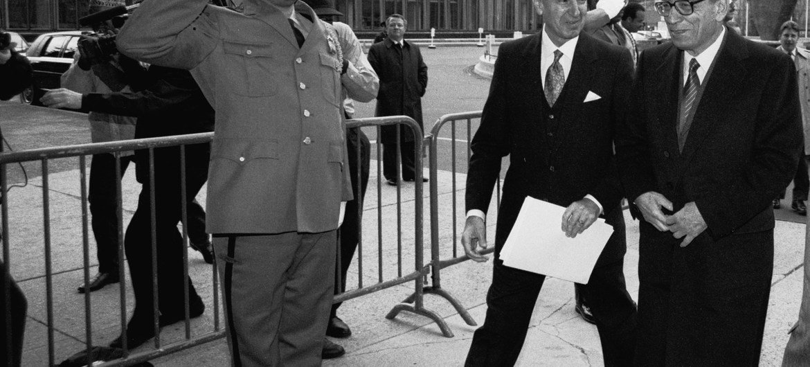 في 2 كانون الثاني عام 1992، بطرس بطرس غالي (من اليمين)، الأمين العام للأمم المتحدة، يصل إلى مدخل الأمانة في أول يوم عمل له في الأمم المتحدة. علي تيمور، رئيس المراسم، يرافقه إلى داخل المبنى. صور الأمم المتحدة / جون إسحاق