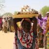 A woman in a red and white headscarf balances a large tray of food on her head in Maiduguri, Nigeria, during a humanitarian aid distribution.