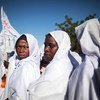 A group of women in white clothing stand together at a protest, holding a flag against a clear blue sky.