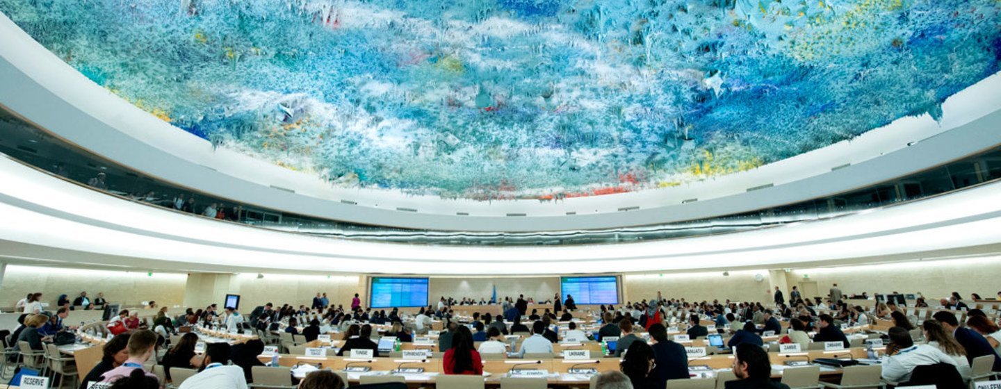 Interior of the United Nations Security Council chamber with delegates seated at tables, each with a nameplate and laptop, under a large, colorful, abstract ceiling artwork.