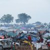 People are gathered among numerous makeshift shelters in a large refugee camp in Wau, South Sudan, with a white car and barbed wire fence in the foreground.