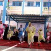 أطفال يرتدون أزياء تقليدية يرقصون على مسرح مفروش بسجادة حمراء في فعالية خارجية، مع وجود لافتات الأونروا والاتحاد الأوروبي في الخلفية.