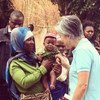 A woman in a headscarf holds a baby who is reaching for a plant while a woman in a blue shirt looks on in a rural village setting.